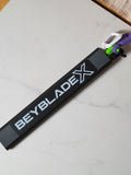 Black three-channel hex rip cord protector with a white Beyblade X logo and white, purple and green cord handles on a pale tabletop.