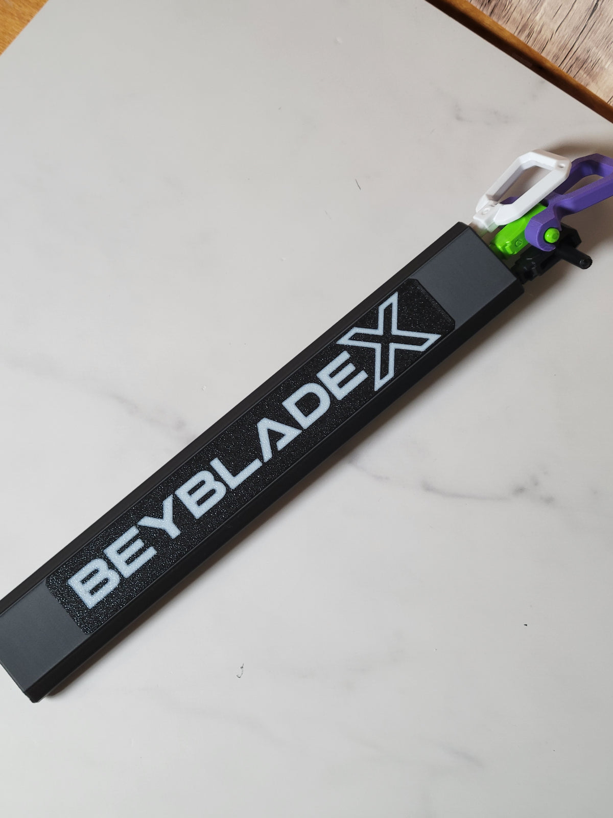 Black three-channel hex rip cord protector with a white Beyblade X logo and white, purple and green cord handles on a pale tabletop.