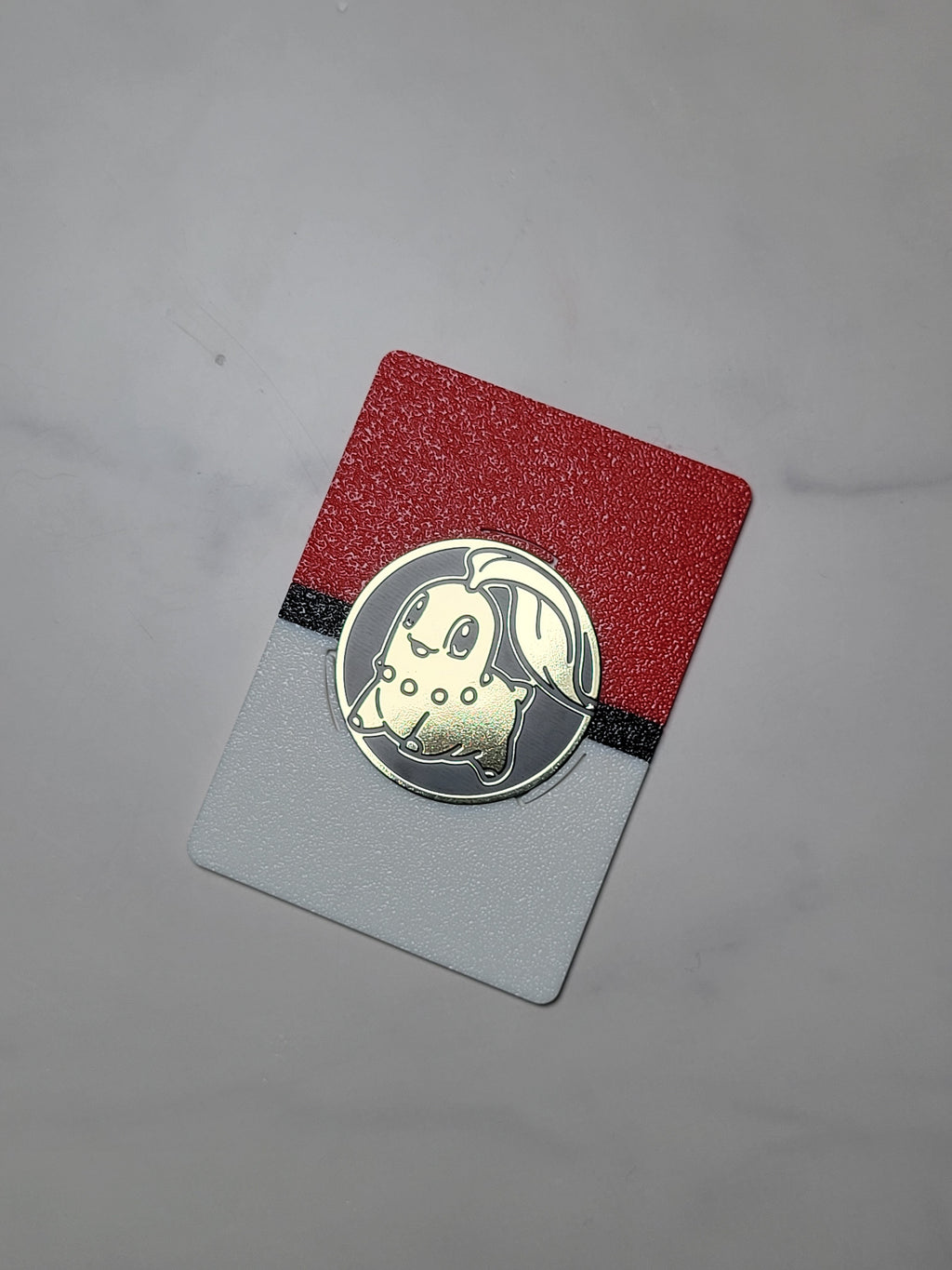 Red, black and white card-size coin display holding a gold Chikorita Pokémon coin on a light marble-patterned tabletop.