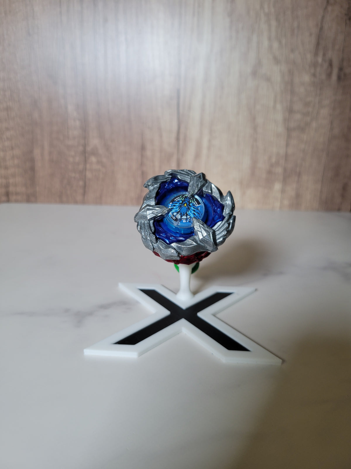 White-and-black X-shaped Beyblade X stand displaying a blue blade above a red ratchet and green bit.