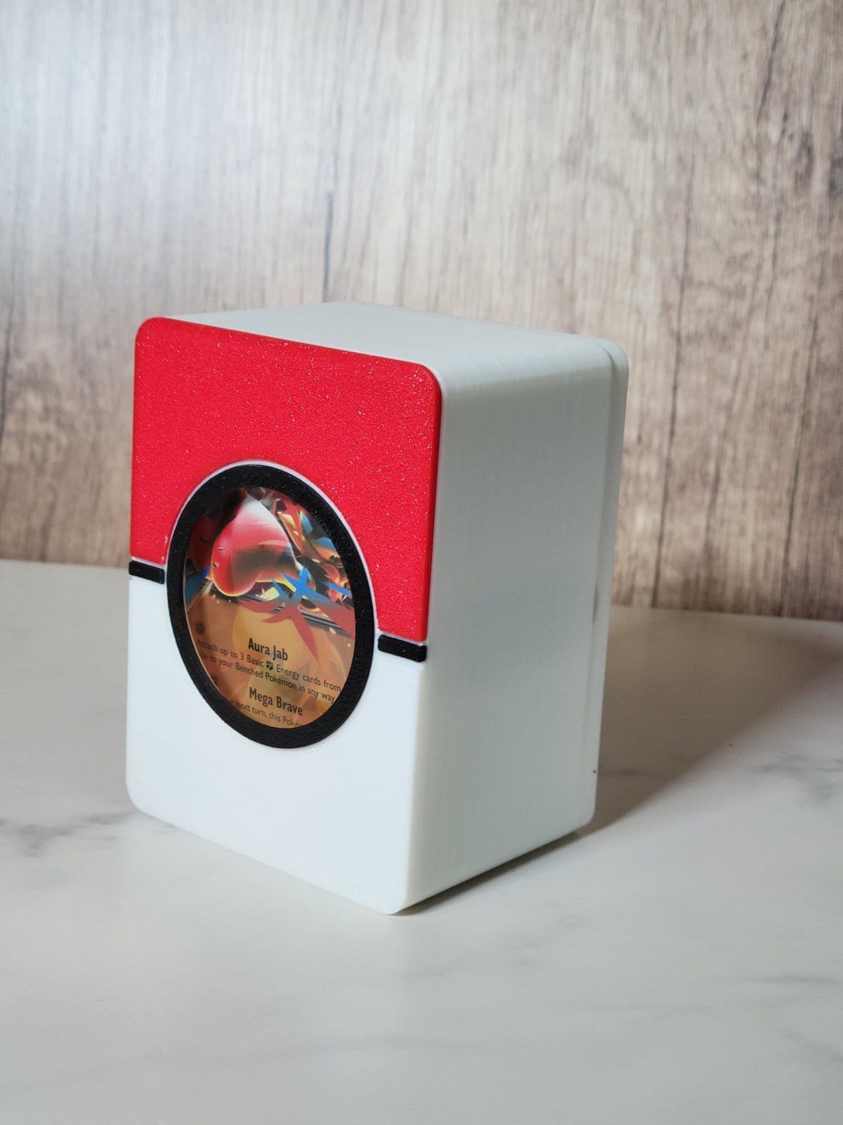 Red, white and black Poké Ball-inspired deck box with a circular front window displaying a sleeved Pokémon card.