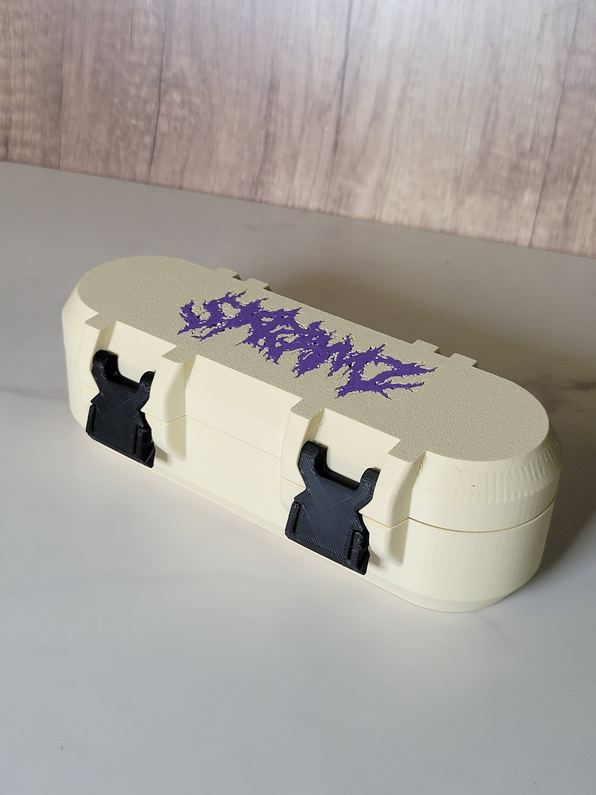 Cream pill-style Beyblade X deck box with purple lid artwork and three black closure clips installed