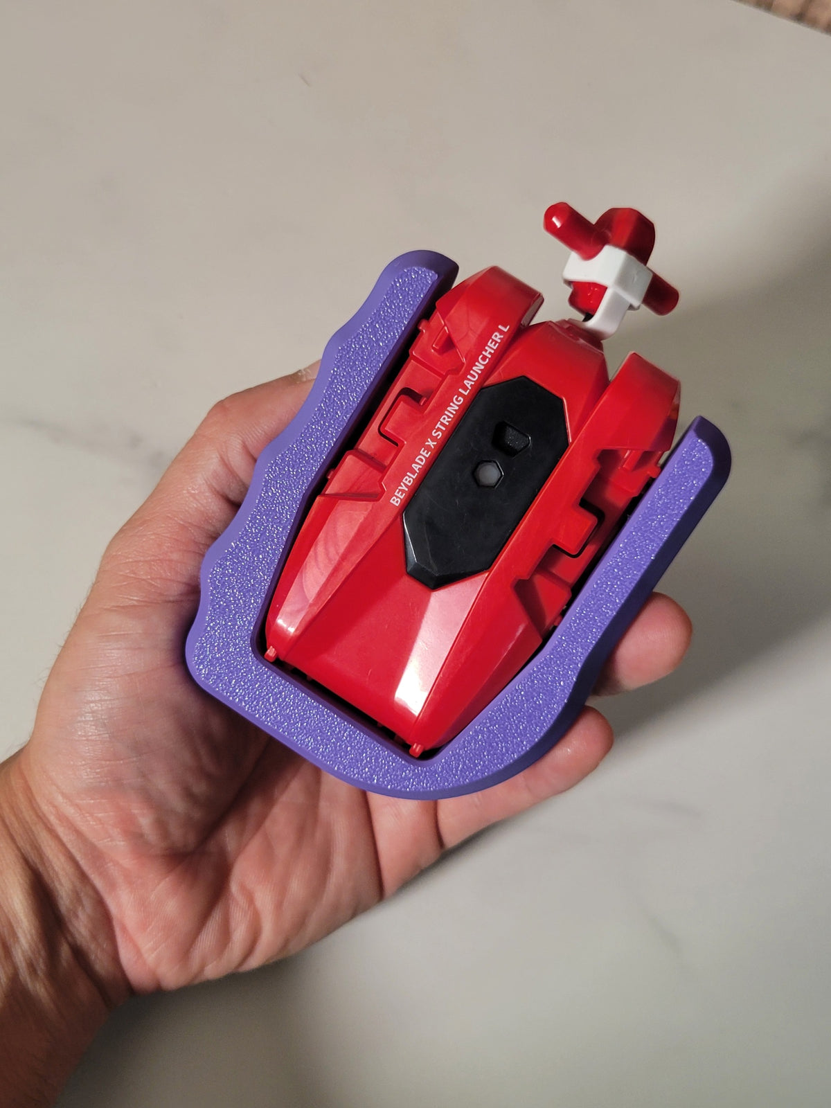 Red Beyblade X string launcher in a purple Ruzz3D grip, held in a hand.